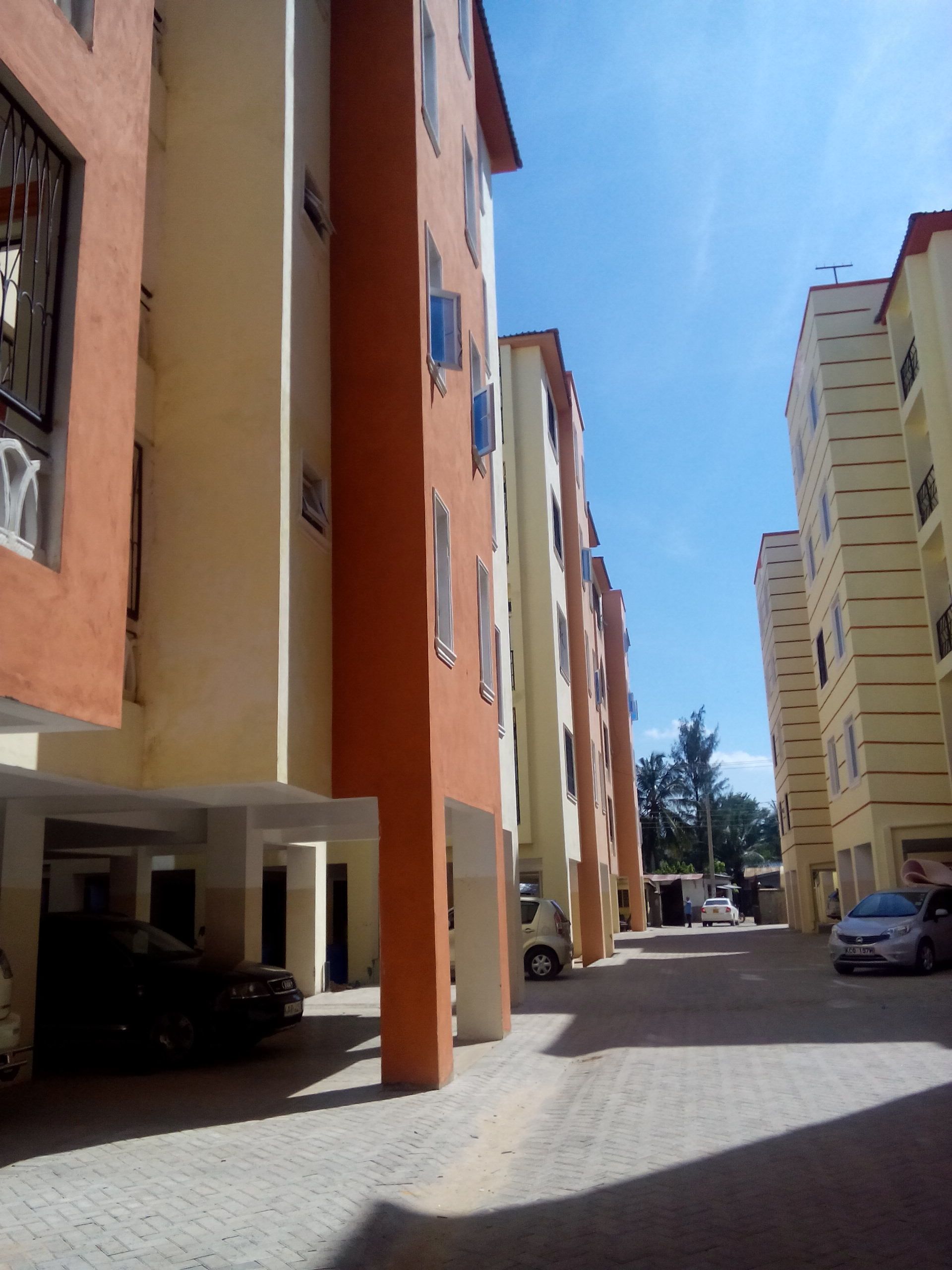 A row of multi-story residential buildings with balconies, a paved walkway, and parked cars.