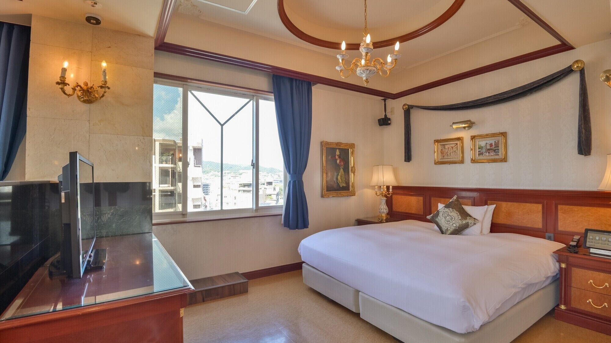 Habitación de hotel con cama, escritorio con televisor, candelabro, ventana con cortinas y obras de arte en la pared.