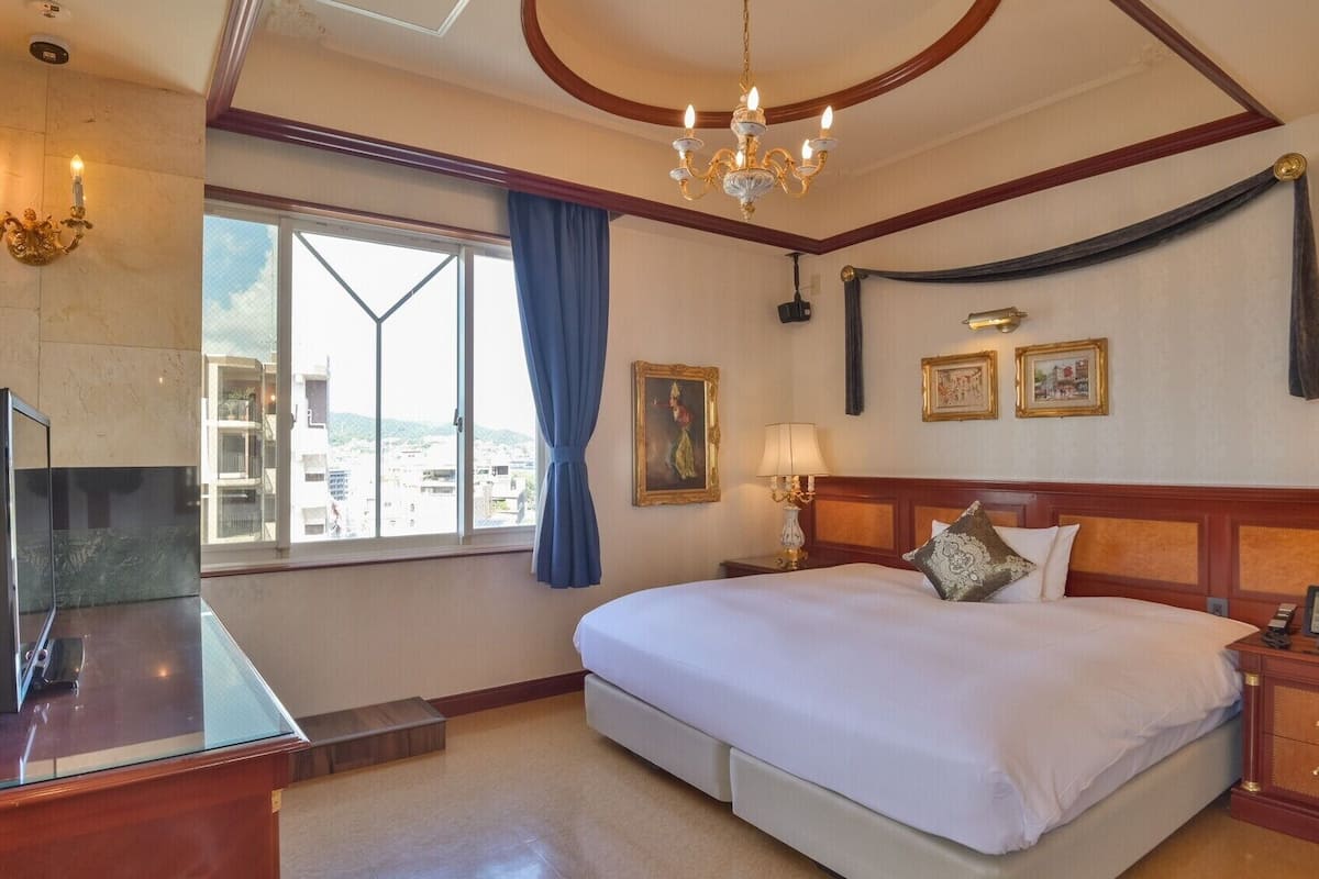 Habitación de hotel con cama, escritorio con televisor, candelabro, ventana con cortinas y obras de arte en la pared.