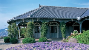 Un bâtiment traditionnel, doté d’un toit en tuiles et de fenêtres en arc de cercle, entouré d’un jardin où poussent des fleurs violettes.