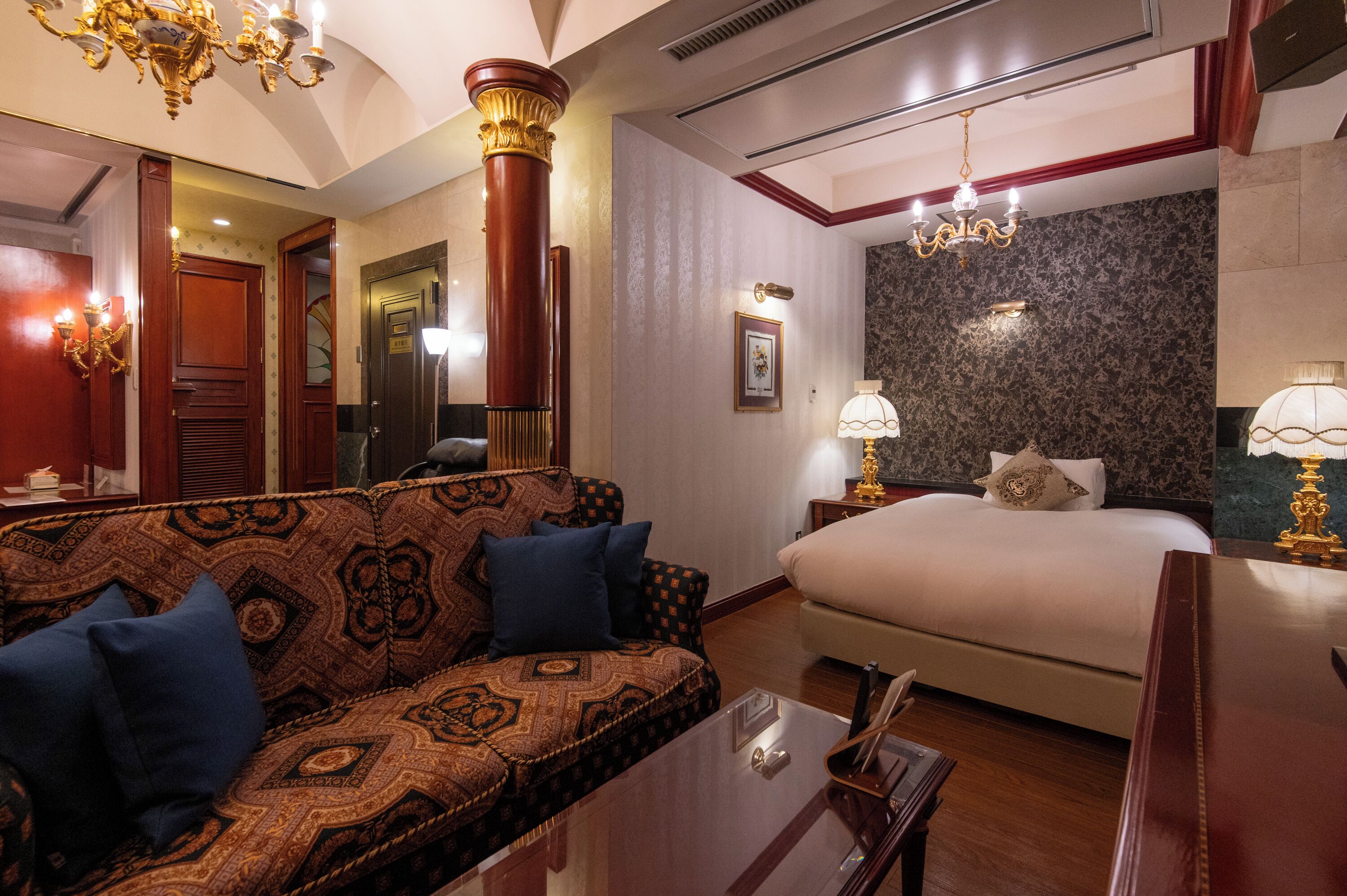 A hotel room with a patterned sofa, a bed, a chandelier, and a large mirror.