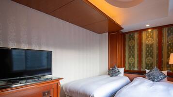 Habitación de hotel con una televisión de pantalla plana sobre un mueble de madera, dos camas con ropa blanca y almohadas estampadas, una pared con diseño verde y techo de madera.