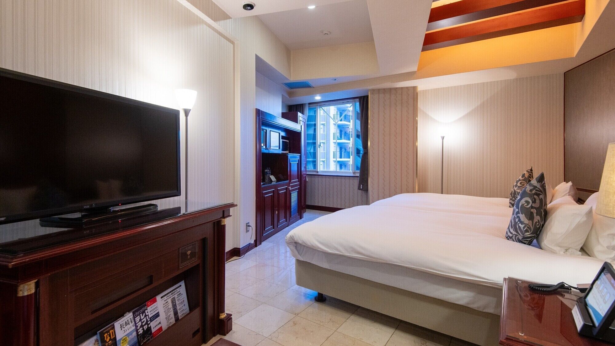 Habitación de hotel con una televisión de pantalla plana grande, una cama con ropa blanca, un escritorio con lámpara y vistas a la ciudad por la ventana.