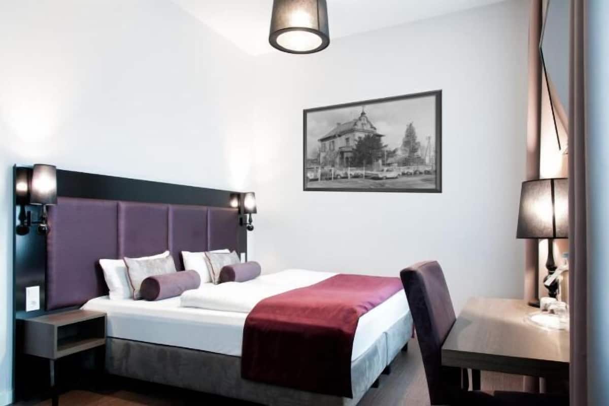 Moderni hotellihuone, jossa on suuri sänky, yöpöytä, tuoli ja työpöytä lampun kanssa.