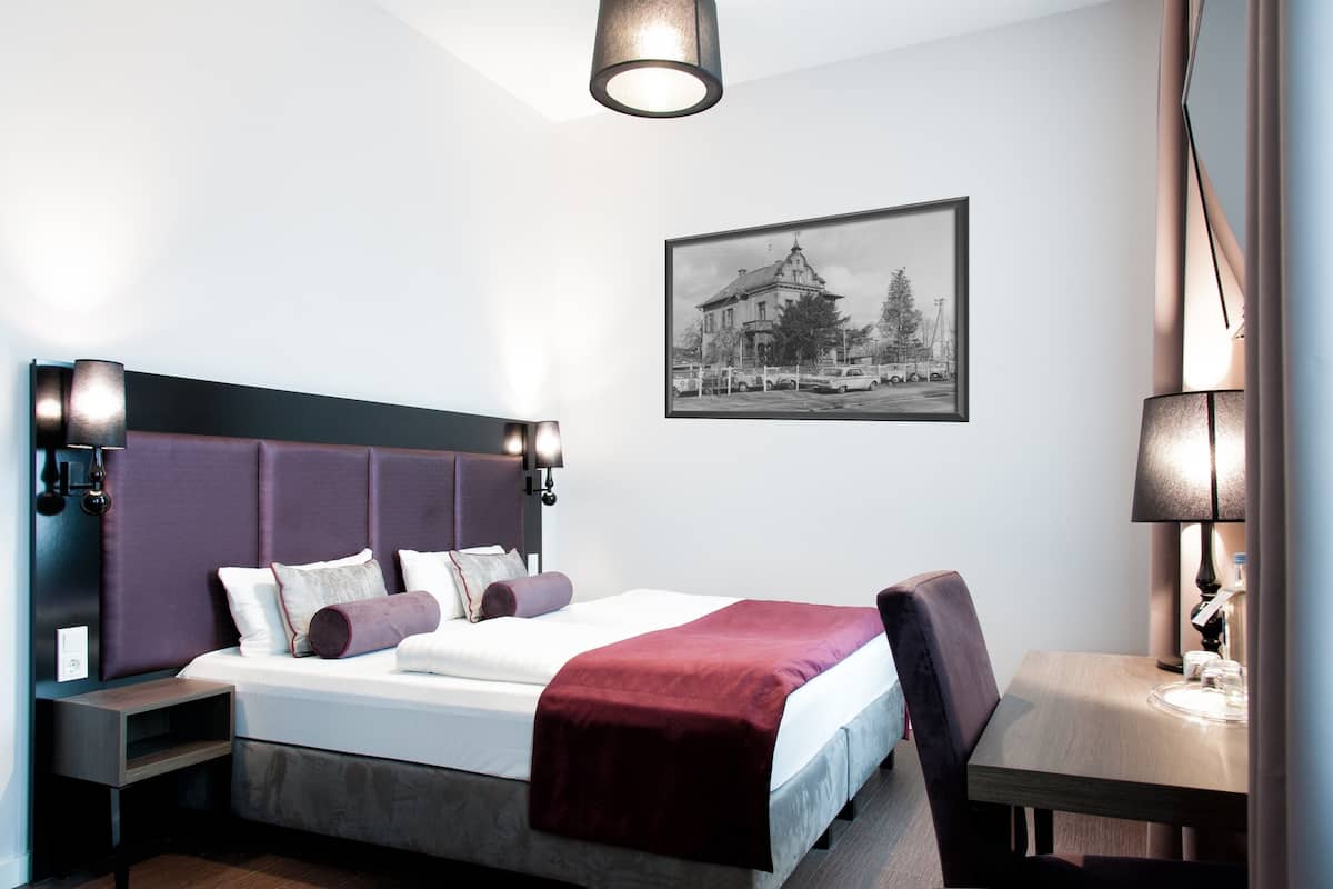 Hotellihuoneessa on suuri sänky, työpöytä ja tuoli. Seinällä on kehystetty kuva ja työpöydällä lamppu.