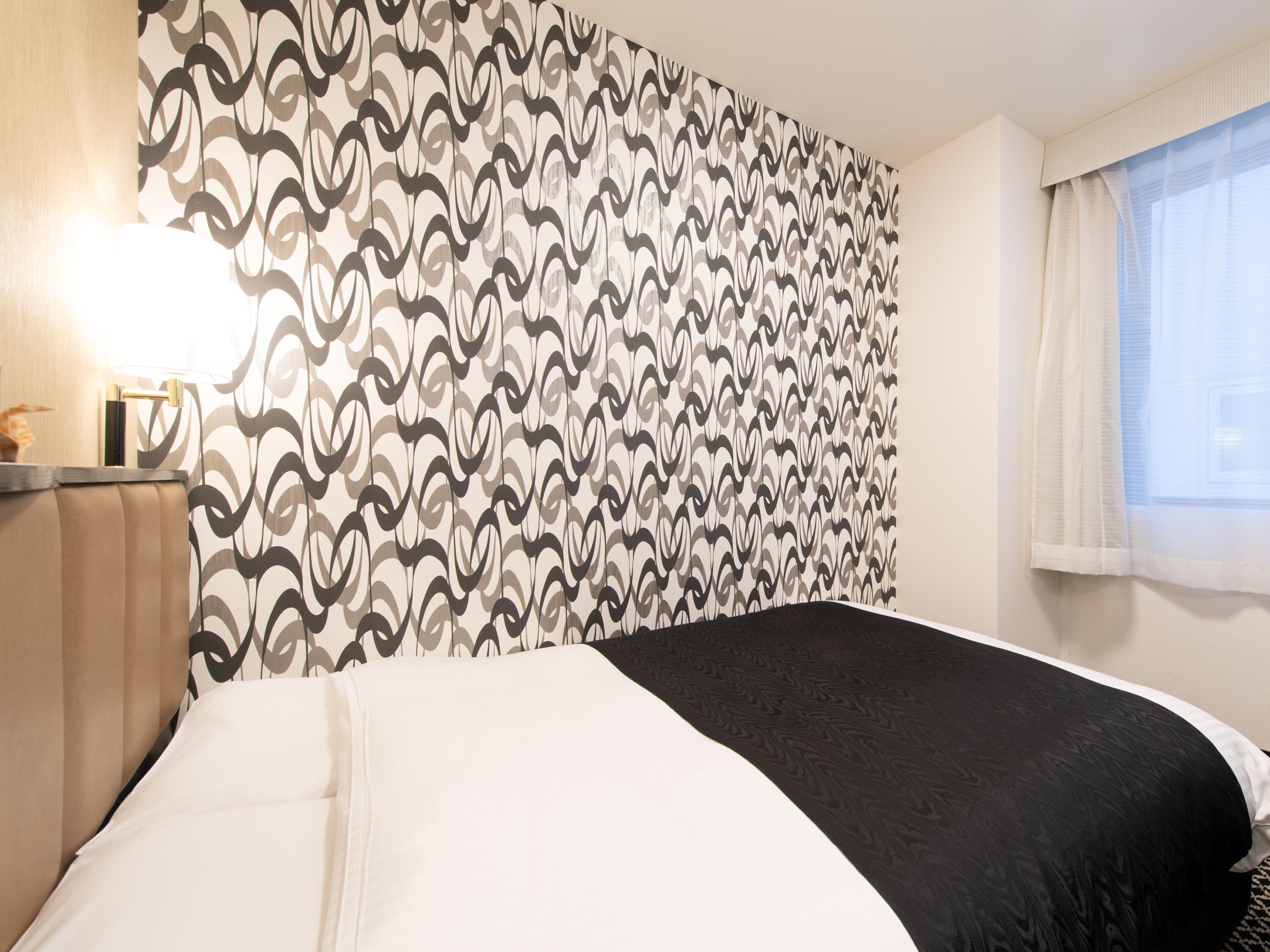 Ein Hotelzimmer mit gemustertem Wandpapier, einem Bett mit weißer und schwarzer Bettwäsche, einem Nachttisch mit Lampe und einem Fenster mit durchsichtigen Vorhängen.