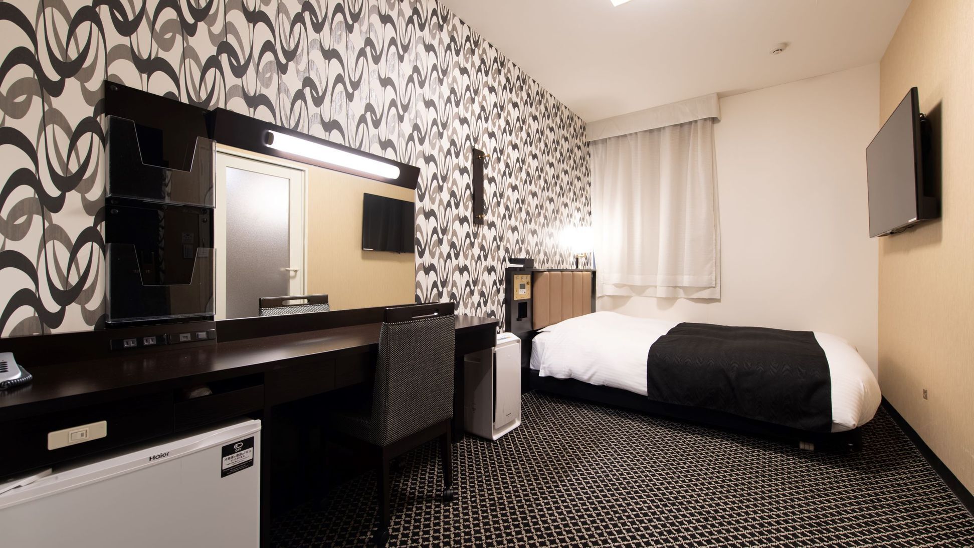 Hotelkamer met een bed, een bureau met stoel, een spiegel, een televisie en een behang met patroon.