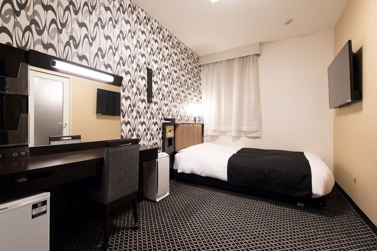 Habitación de hotel con una cama, un escritorio con silla, un espejo, un televisor y un papel tapiz decorado.