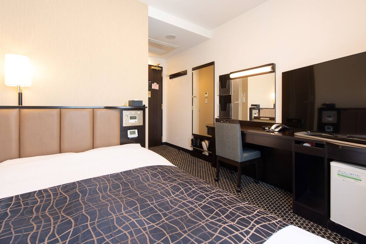 Habitación de hotel con una cama grande, un escritorio con silla, un espejo, un refrigerador pequeño y un panel de control en la pared.