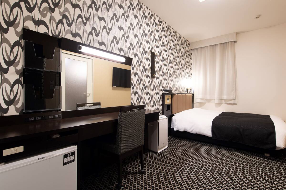 Habitación de hotel con cama, escritorio, silla, espejo, televisor y panel de control en la pared.