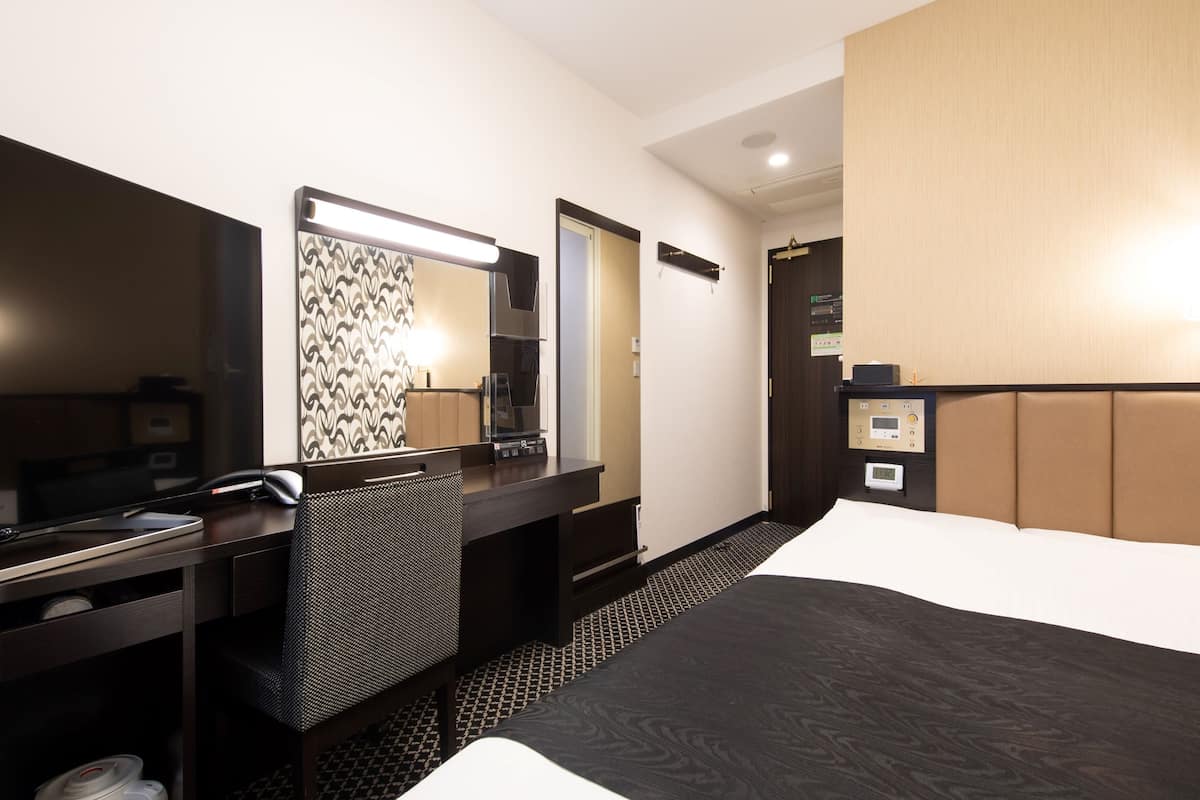 A hotel room with a bed, a desk with a chair, a large mirror, a bedside table, and a control panel.