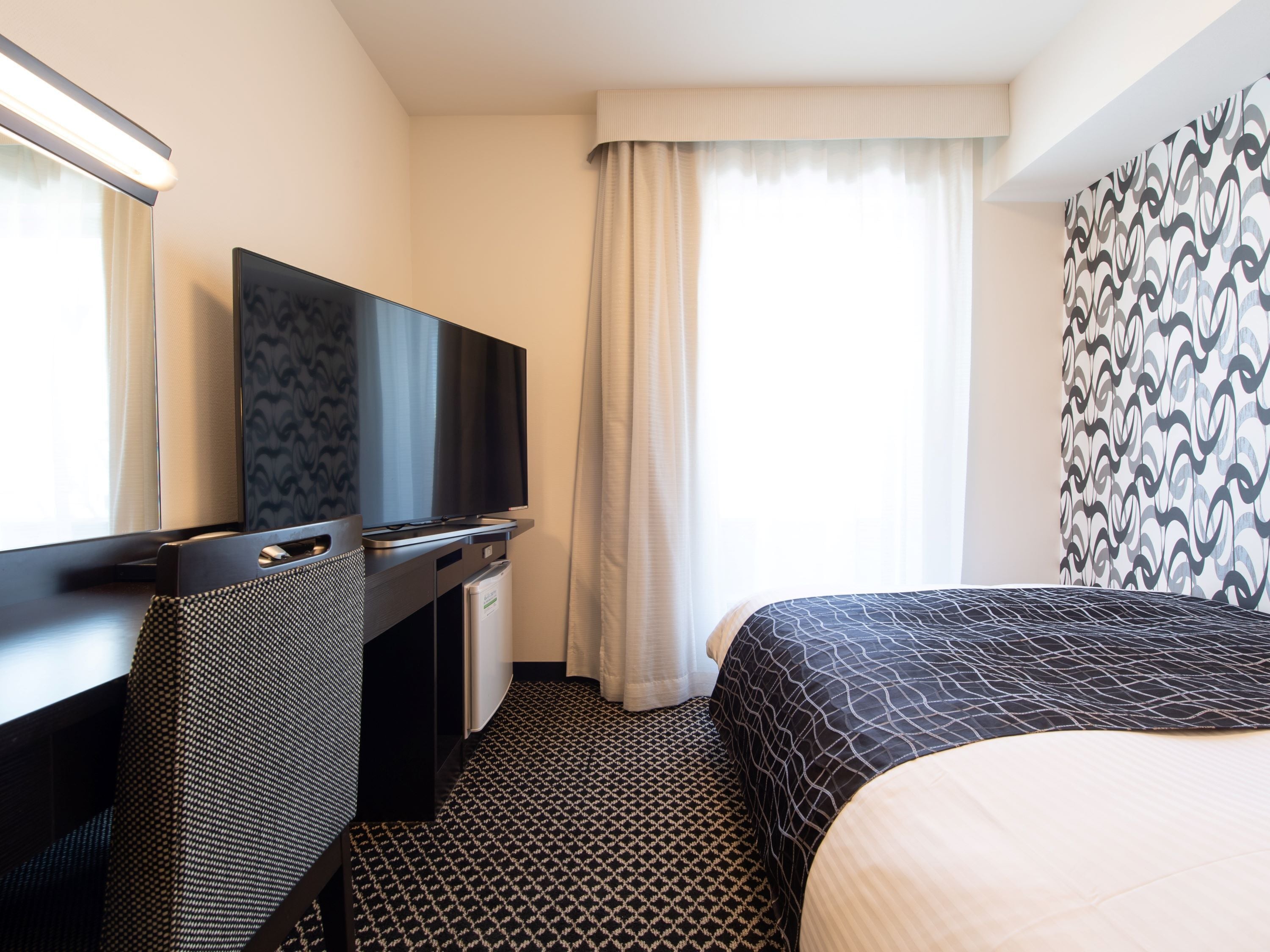 A hotel room with a bed, a desk with a flat-screen TV, a minibar, and a window with curtains.
