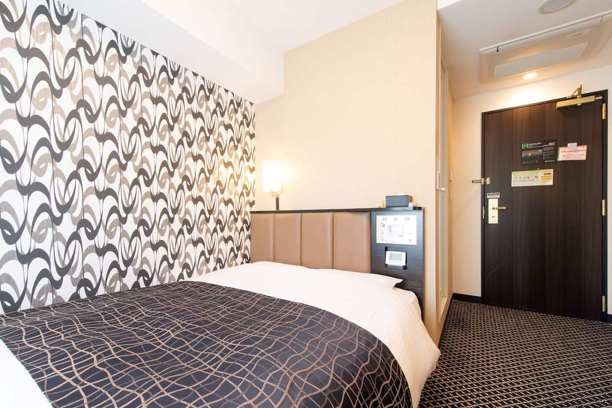 A hotel room with a bed, a control panel, a door, and a patterned wall.
