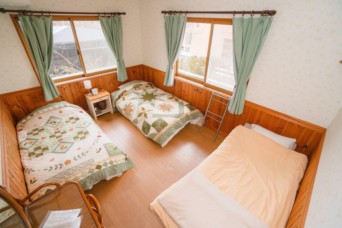 A room with two beds, a chair, a small table with a lamp, and a window with curtains.