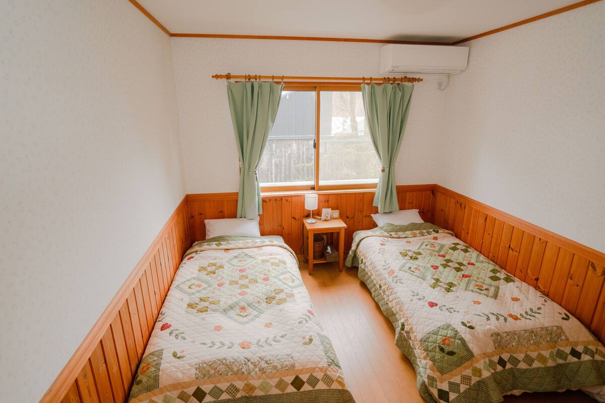 A room with two wooden beds, a small wooden nightstand, a window with curtains, and an air conditioning unit.