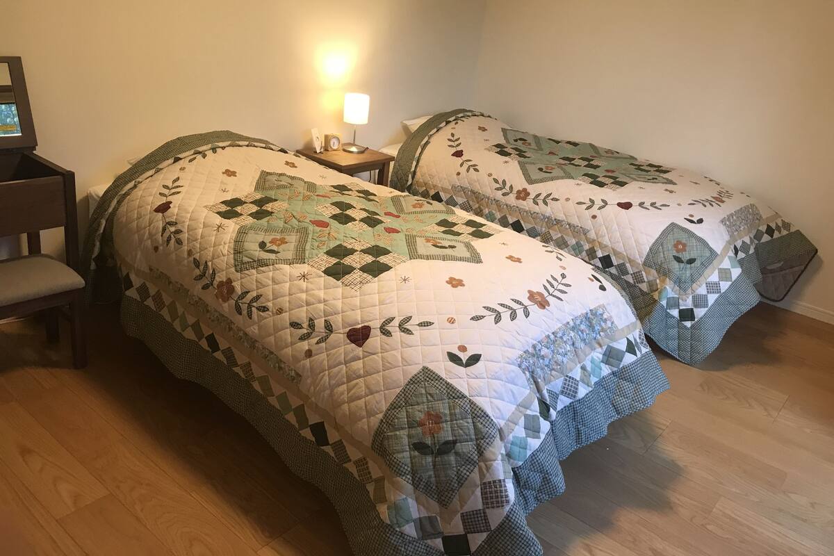 Two beds with quilts, a bedside table, a lamp, and a chair in a room with wooden flooring.
