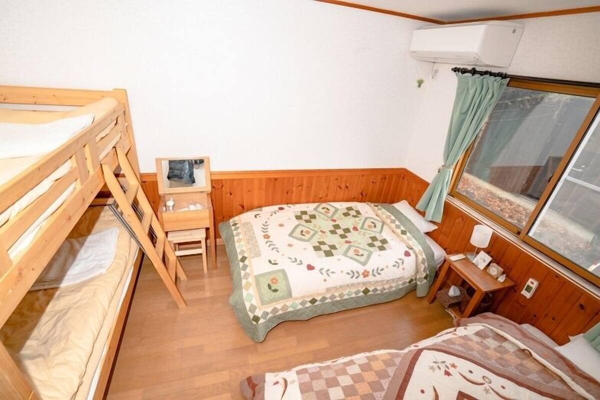A small, cozy room with a bunk bed, a desk, a chair, a bed with a patterned blanket, and a window with curtains.