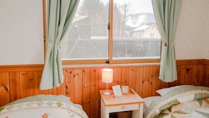 A bedroom with a wooden paneled wall, a window with curtains, a bedside table with a lamp, and a bed with a quilt. - Guesthouse Chaconne Karuizawa (Karuizawa)