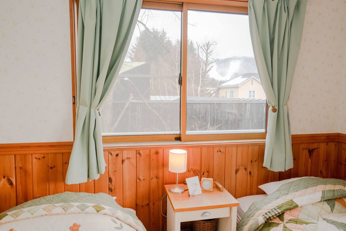 A bedroom with a wooden paneled wall, a window with curtains, a bedside table with a lamp, and a bed with a quilt.