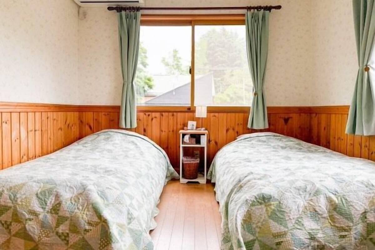 A room with two beds, wooden walls, a window with curtains, and a small bedside table with a lamp.