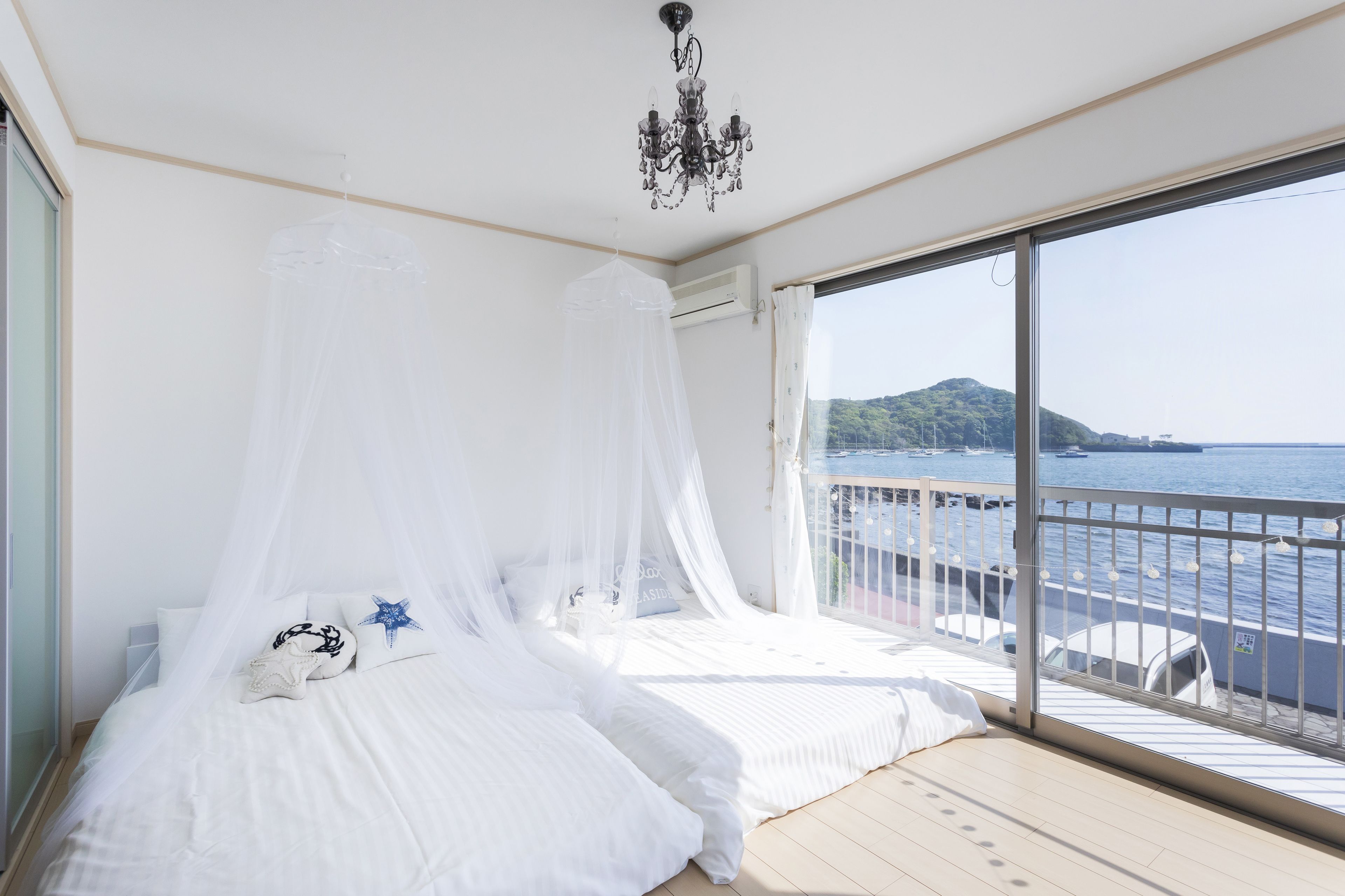 A bedroom with a bed under a white canopy, a chandelier, and a view of the sea through a large window.