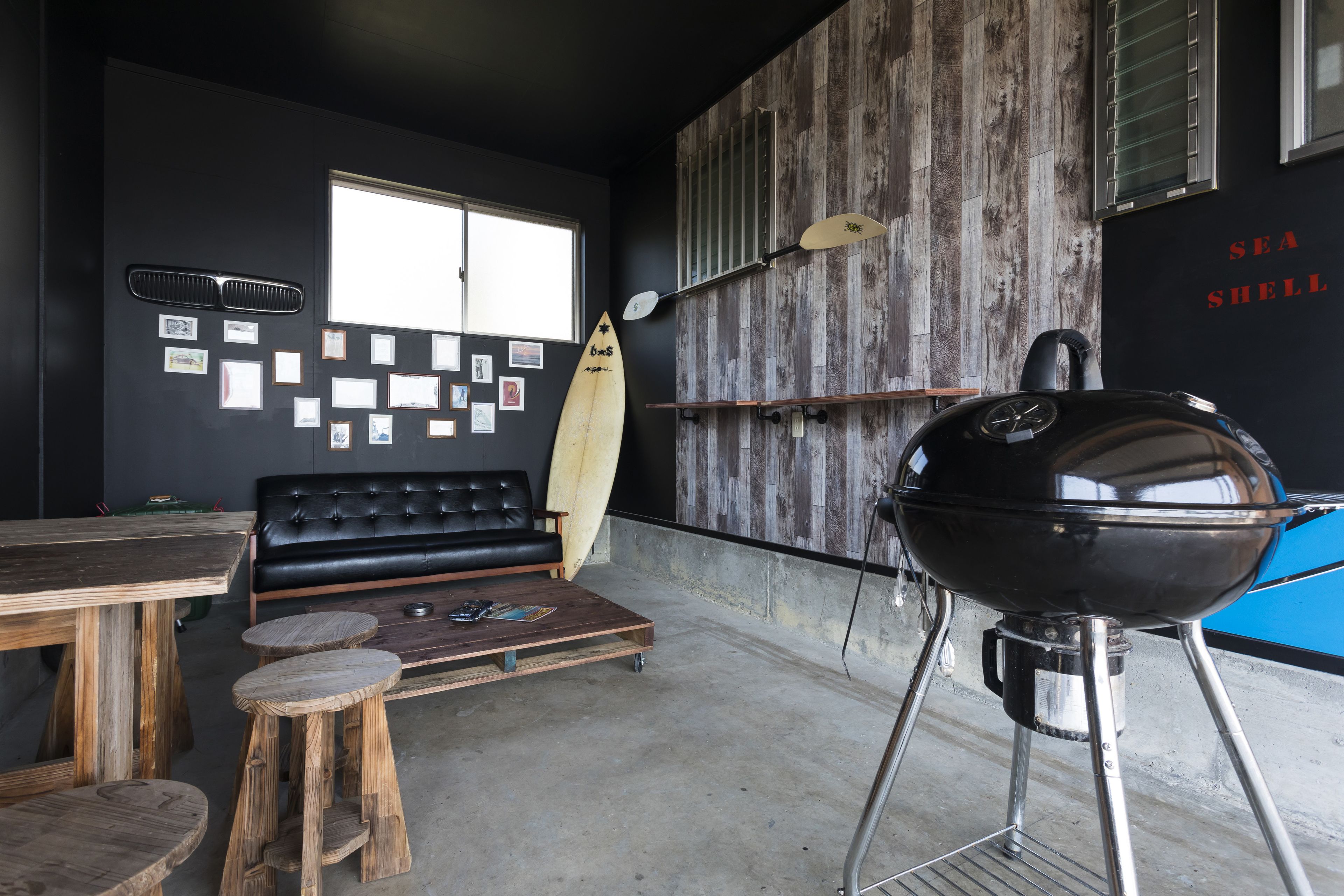 A modern interior with a black leather couch, wooden stools, a barbecue grill, and a surfboard on the wall.