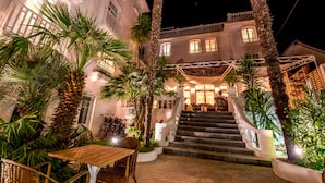 Un edificio bien iluminado con una escalera que conduce a un balcón, rodeado de palmeras y mobiliario exterior.