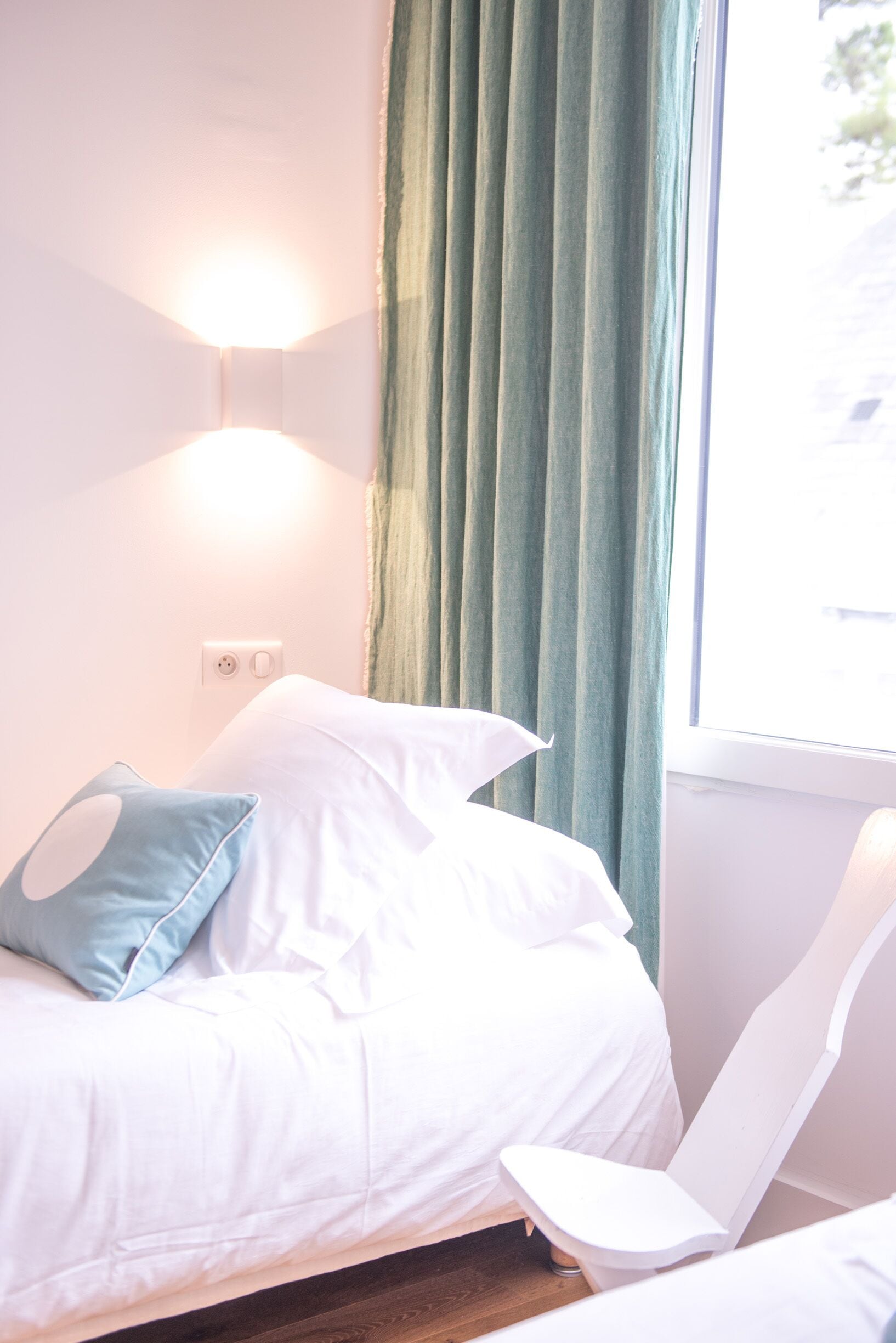 A neatly made bed with white linens, a blue pillow, and a white chair beside it.