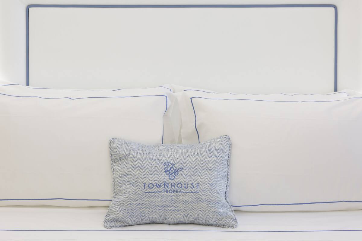 A bed with white bedding and blue trim, a pillow with embroidered text, and a blue and white patterned pillow.