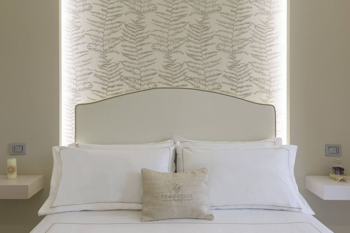 A neatly made bed with white linens and a single decorative pillow, a headboard with a leaf pattern, and a window with a leaf-patterned curtain.