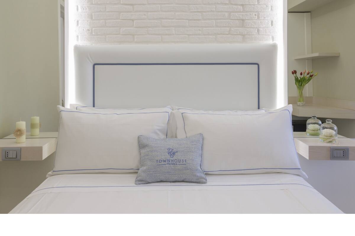 A bedroom with a white brick headboard, a bed with white bedding, a nightstand with a candle, and a vase with flowers on the nightstand.