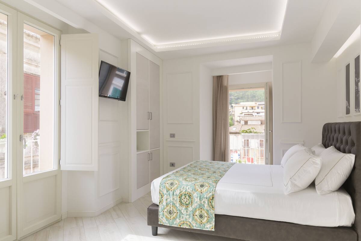 A bedroom with a bed, a TV mounted on the wall, and a window with a view of buildings.