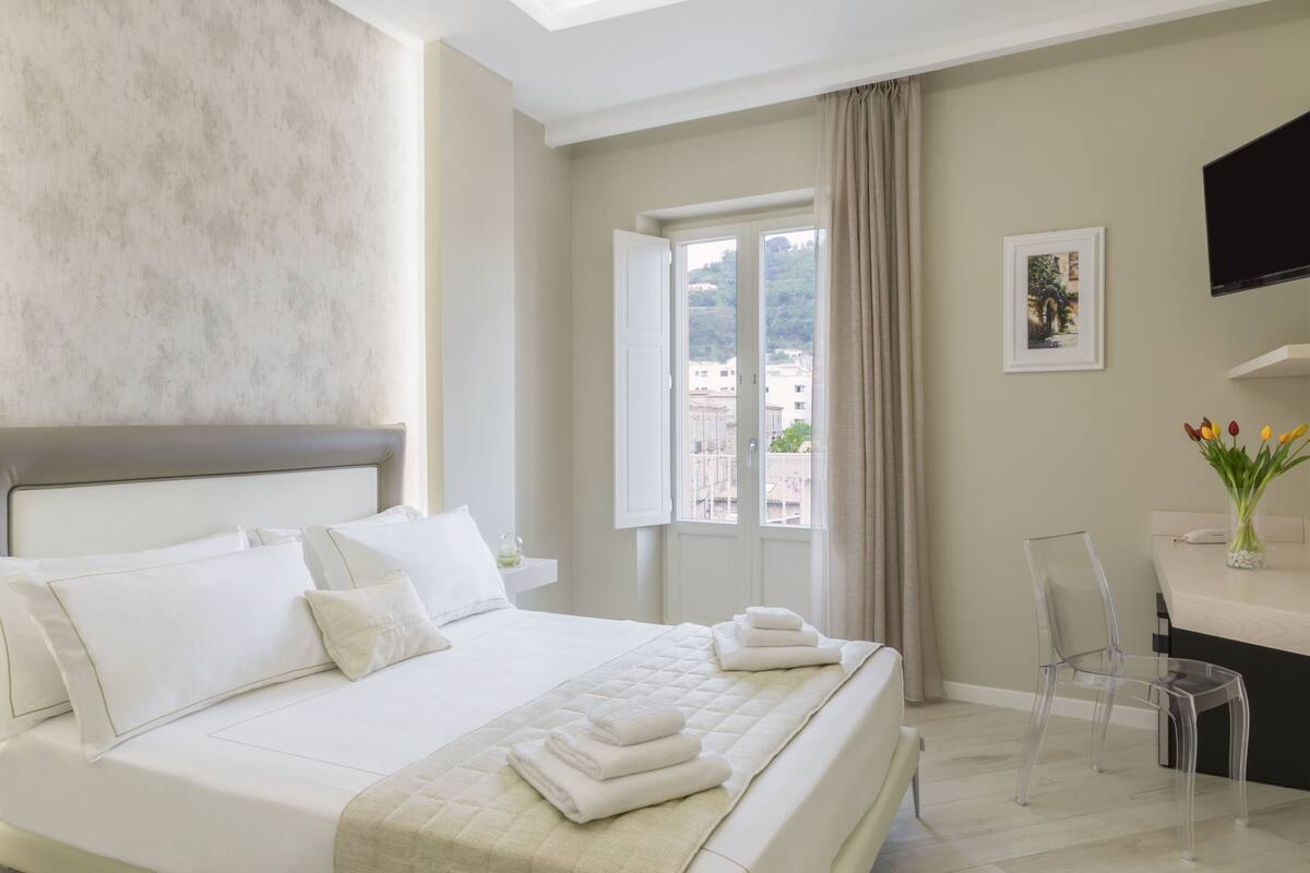 A hotel room with a large bed, a clear glass chair, a flat-screen TV, and a window with a view of buildings.