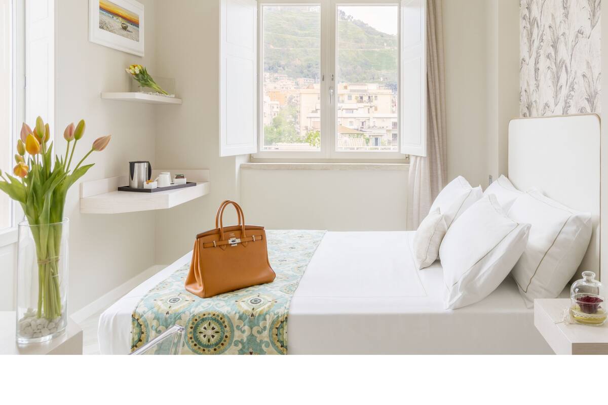 A bedroom with a bed, a window with a view of buildings, a vase with flowers, and a handbag on the bed.