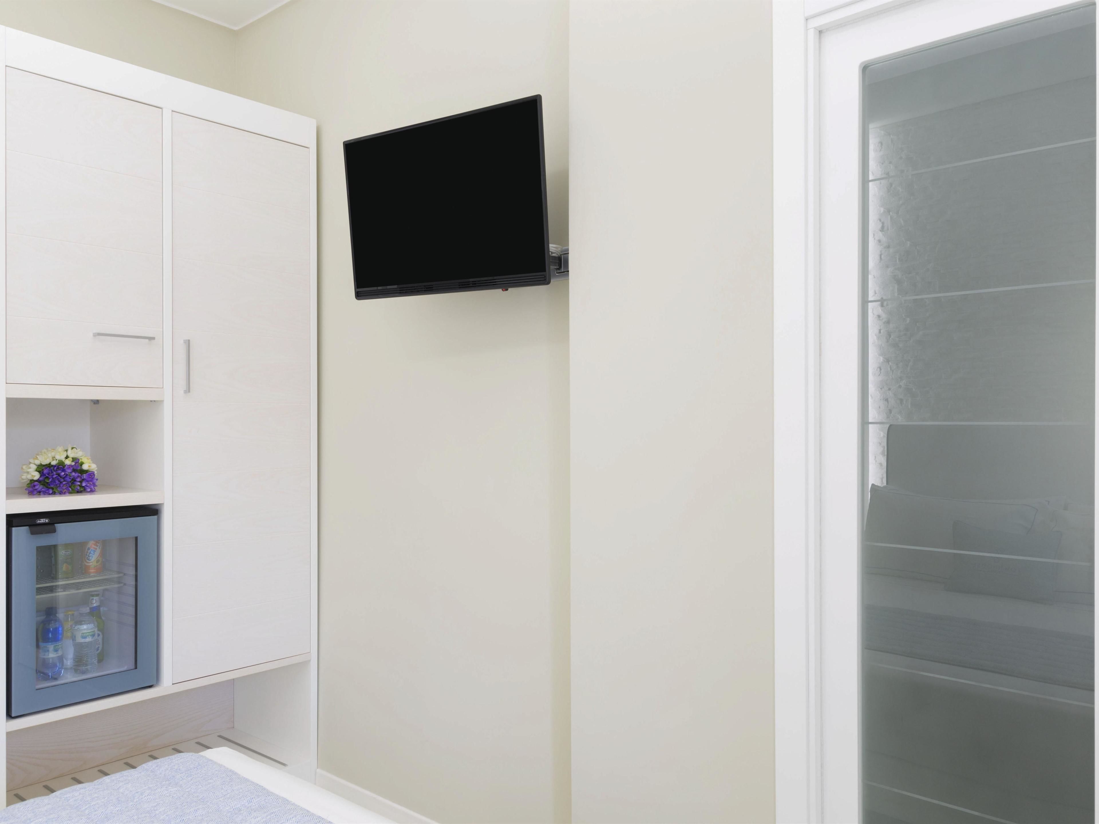 A room with a white wardrobe, a small fridge, a mounted TV, and a glass shower enclosure.