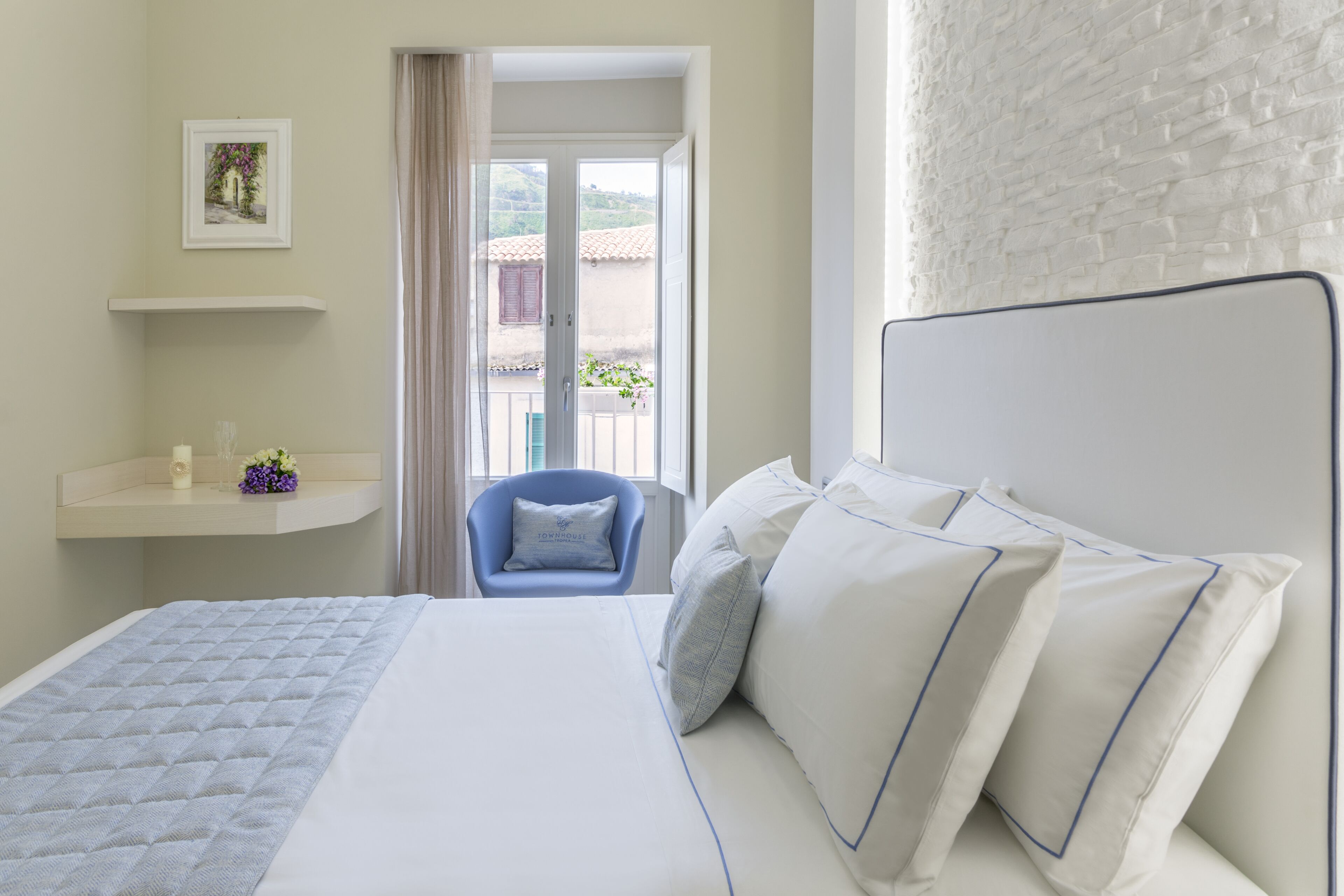 Une chambre à coucher avec un lit, une chaise, une étagère avec un vase et une vue sur l’extérieur.