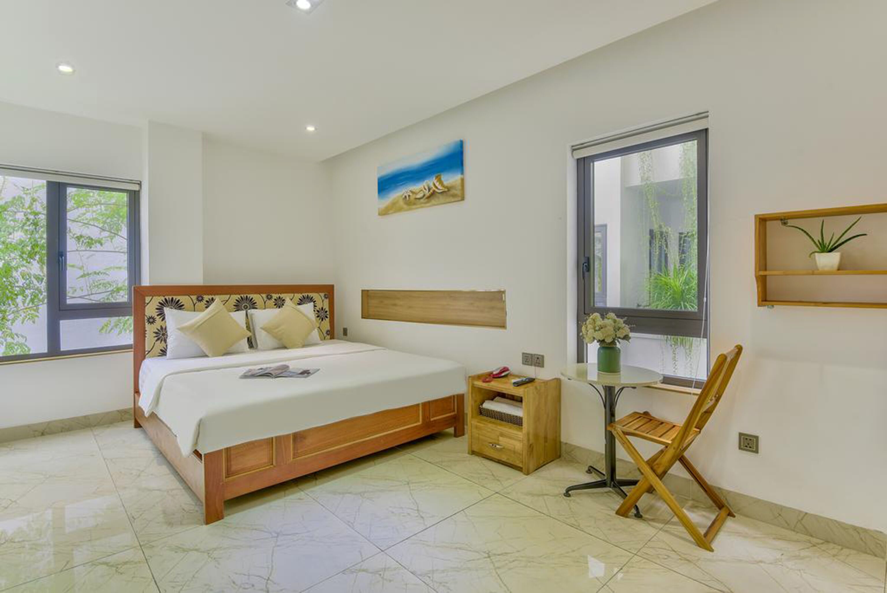 A bedroom with a bed, a small table with a plant, a chair, and a painting on the wall.