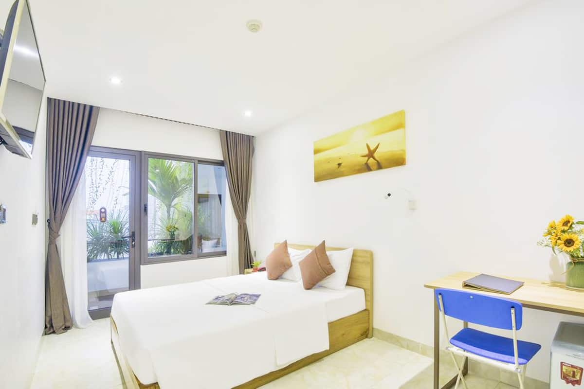 A hotel room with a bed, a desk, and a chair. There is a window with curtains, a painting on the wall, and a vase with flowers on the desk.
