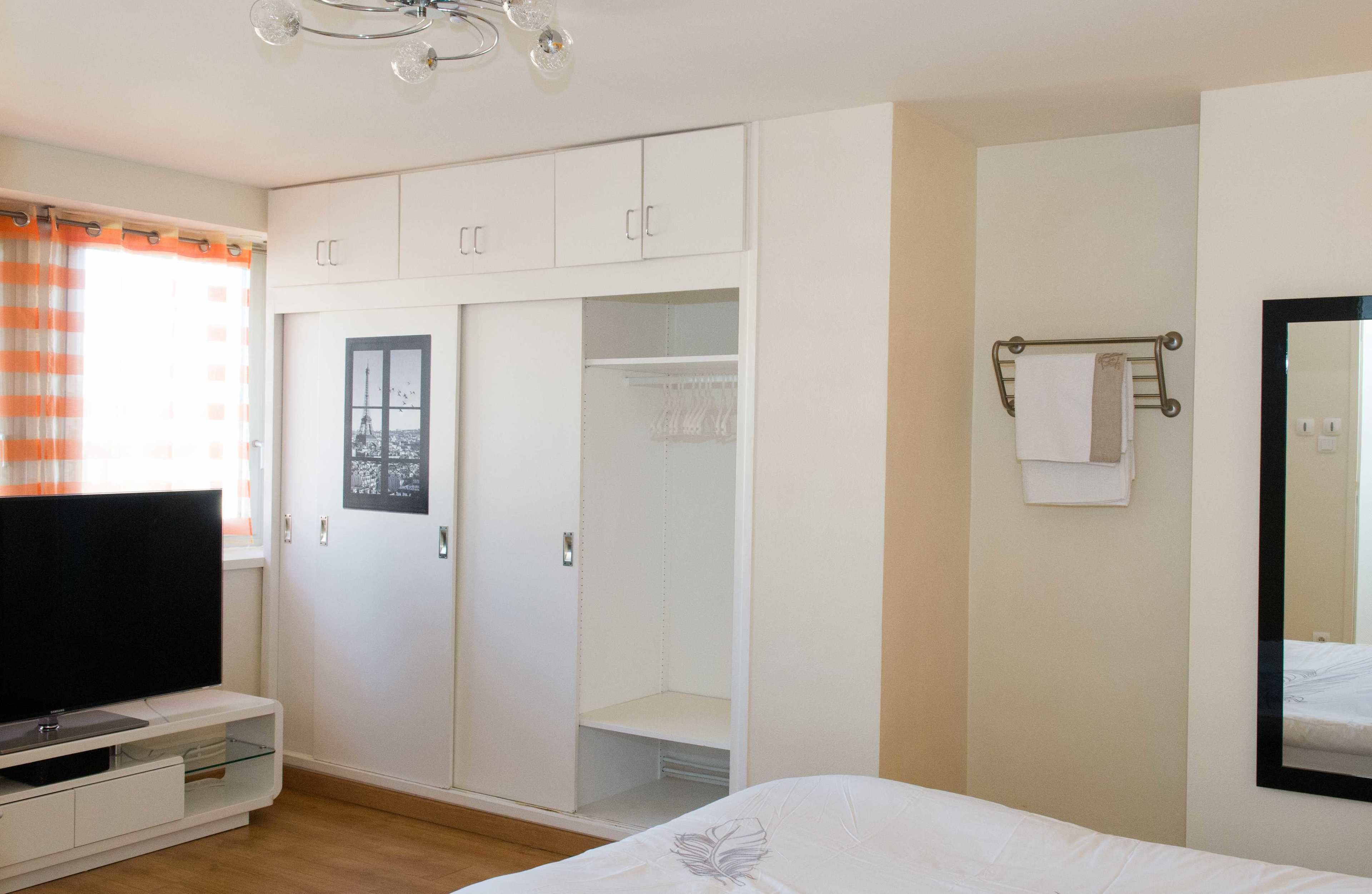 A bedroom with a bed, a TV on a stand, a wardrobe, and a bathroom with a mirror and towel rack.