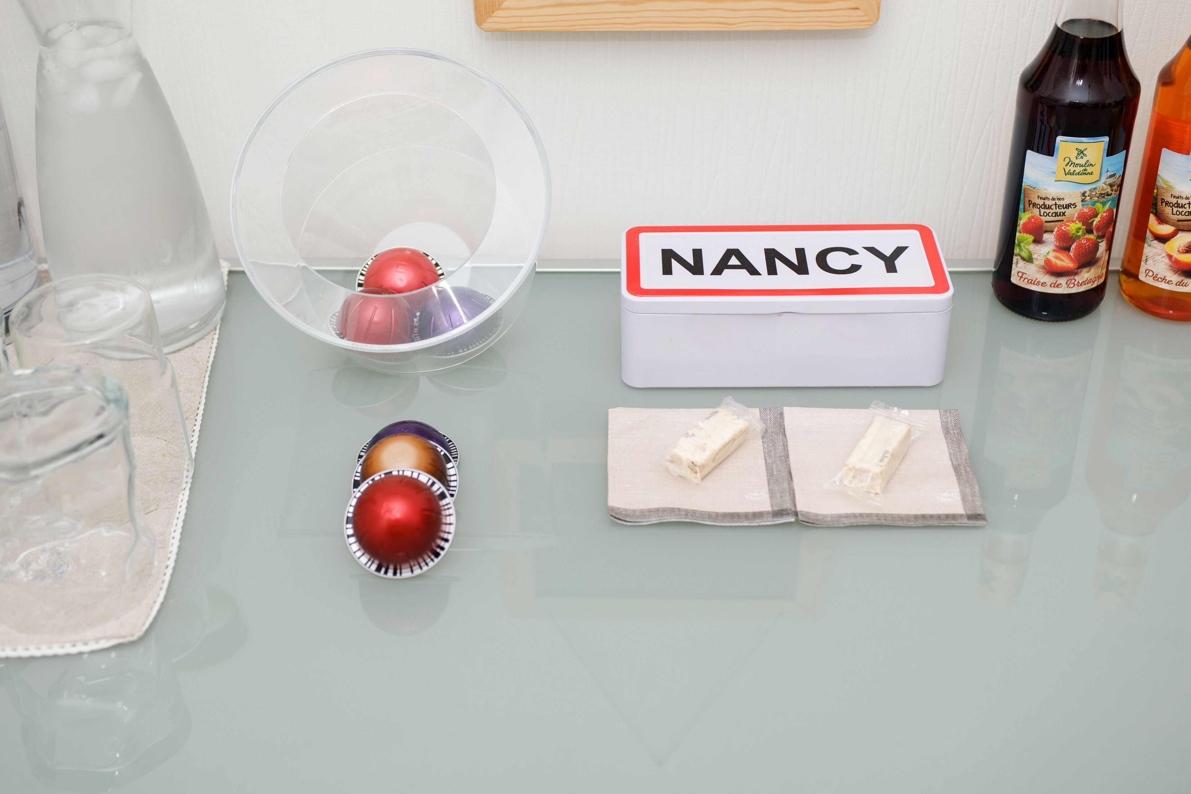 Ein Tisch mit einer klaren Plastikschale, einer weißen Schachtel mit dem Aufdruck 'NANCY', einem kleinen roten Gegenstand und zwei Päckchen weißem Pulver.