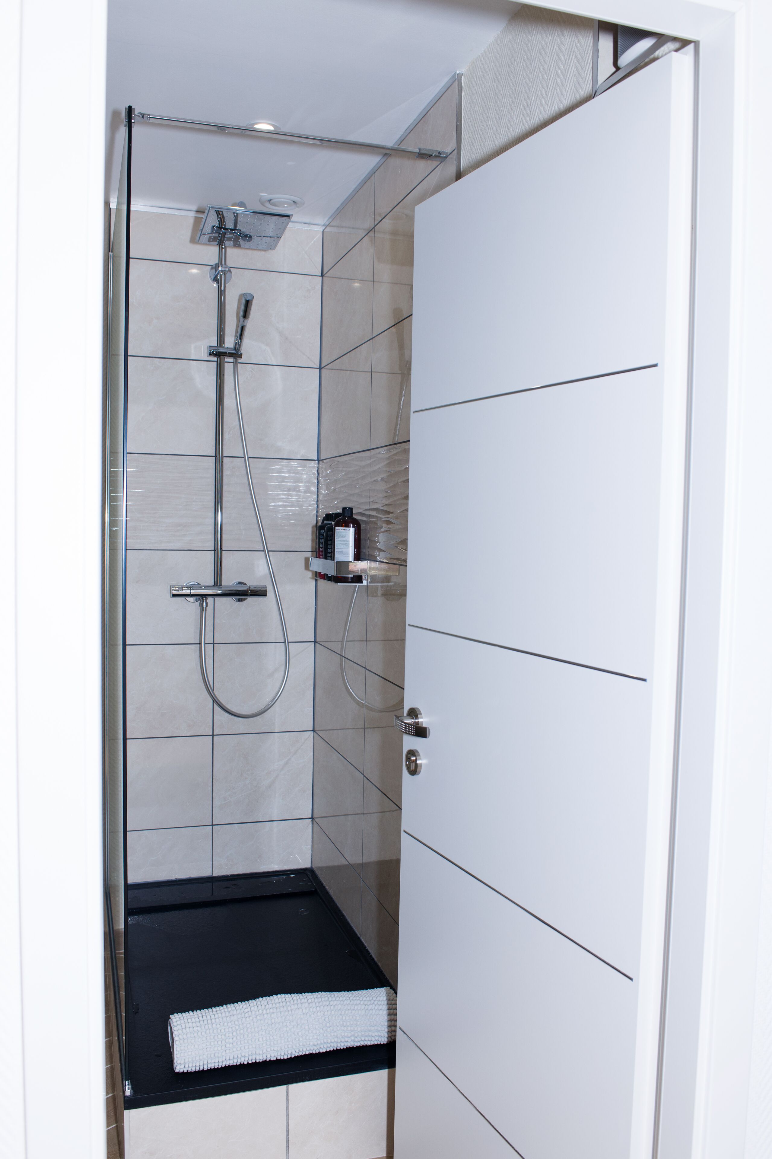 Une cabine de douche moderne avec une porte en verre, un tapis noir et une petite étagère contenant des articles de toilette.