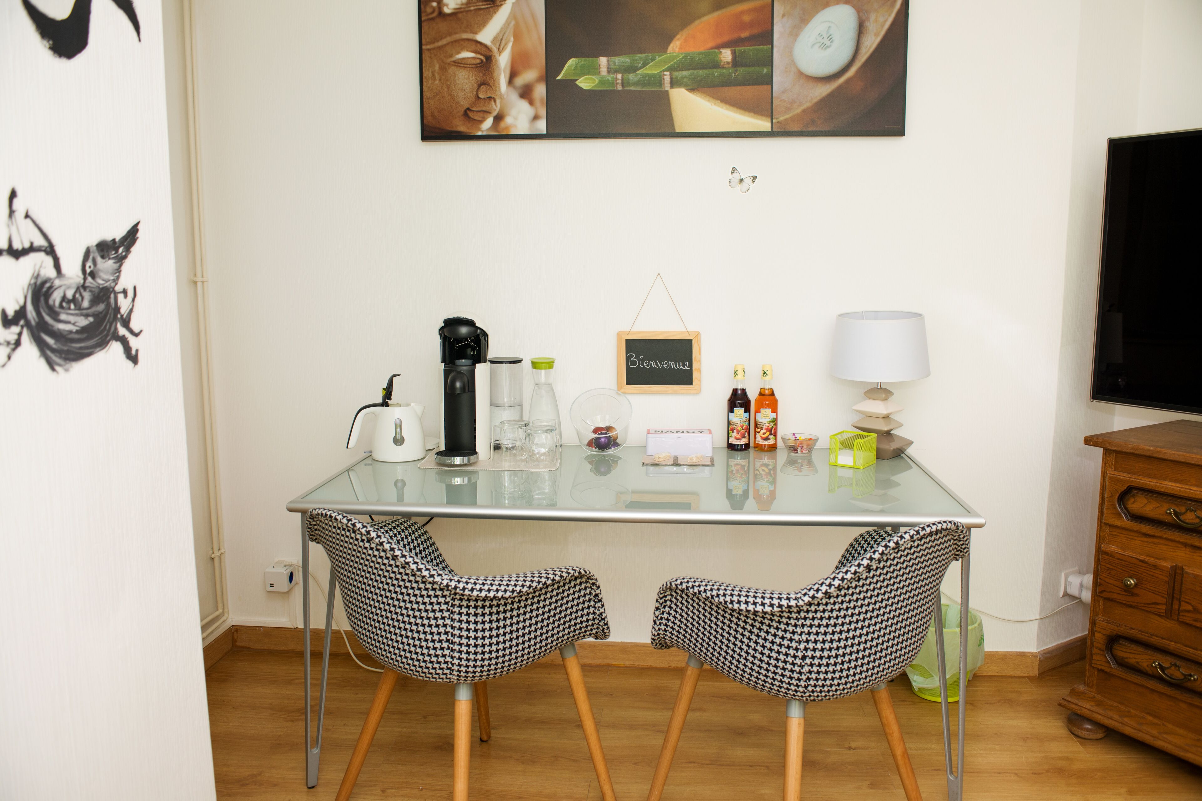 Ein kleiner, moderner Raum mit einem Glastisch, zwei gemusterten Stühlen, einer Kaffeemaschine, einer Lampe und einem Fernseher.
