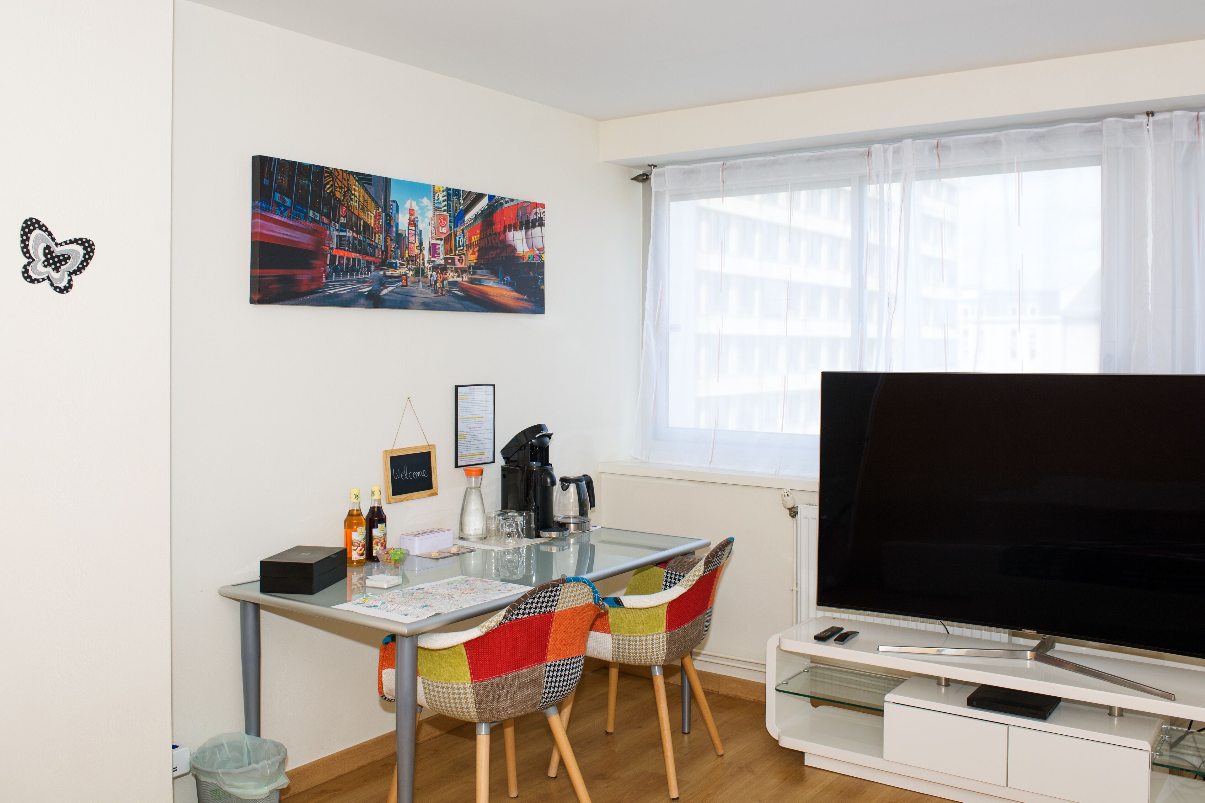 A modern living room with a glass dining table, colorful chairs, a flat-screen TV, and a cityscape painting on the wall.