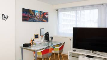 A modern living room with a glass dining table, colorful chairs, a flat-screen TV, and a cityscape painting on the wall.