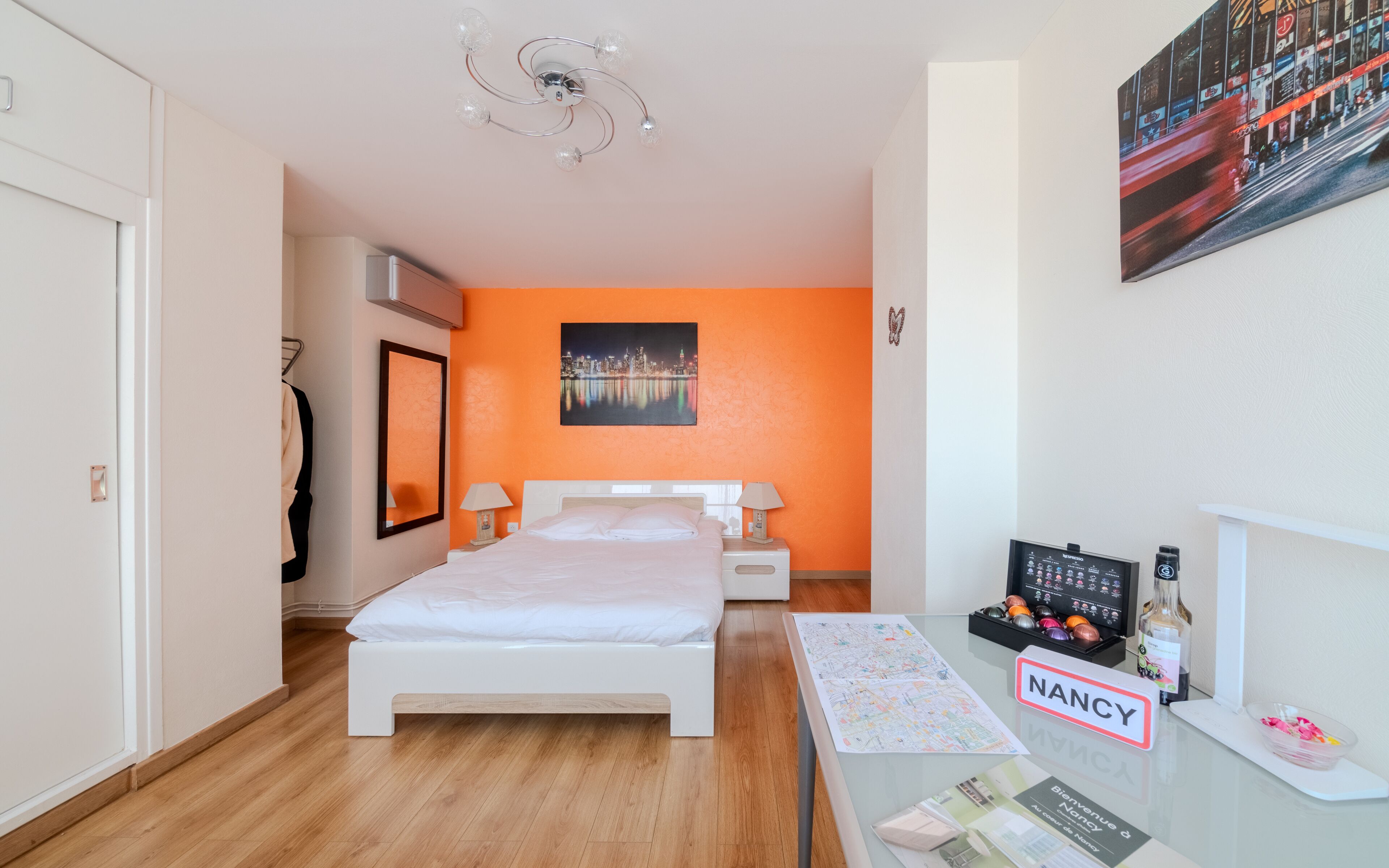 A hotel room with a bed, a nightstand, a desk with a computer, a chair, a coat rack, a mirror, and a cityscape painting on the wall.