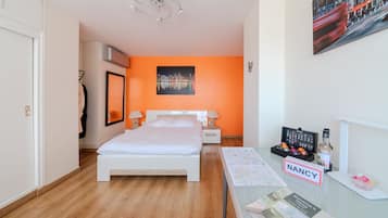 A hotel room with a bed, a nightstand, a desk with a computer, a chair, a coat rack, a mirror, and a cityscape painting on the wall.