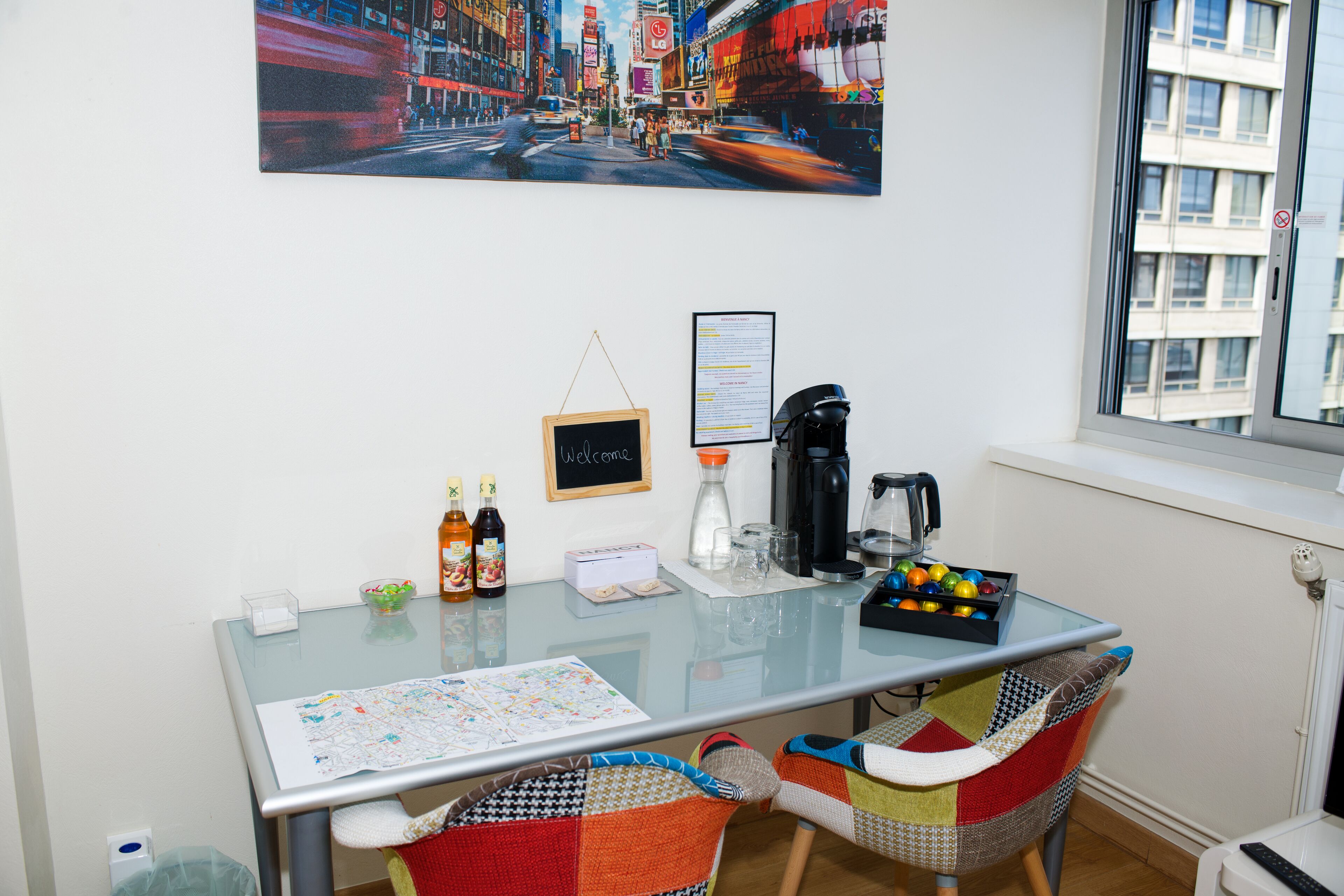 A room with a glass table, two chairs, a coffee maker, a small chalkboard, and a window with a view of buildings.