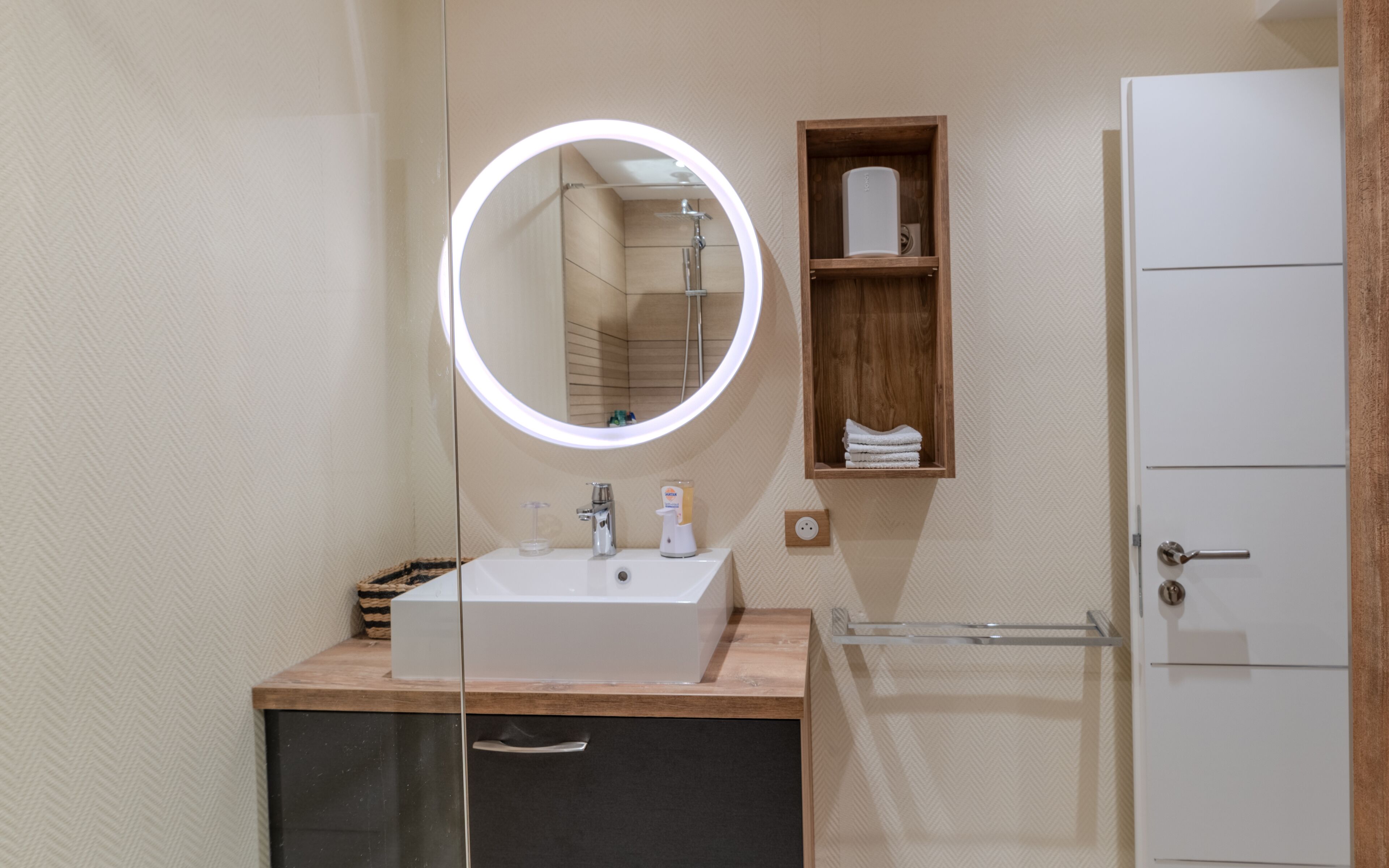 A modern bathroom with a round mirror, a sink, a wooden shelf with towels, and a cabinet.