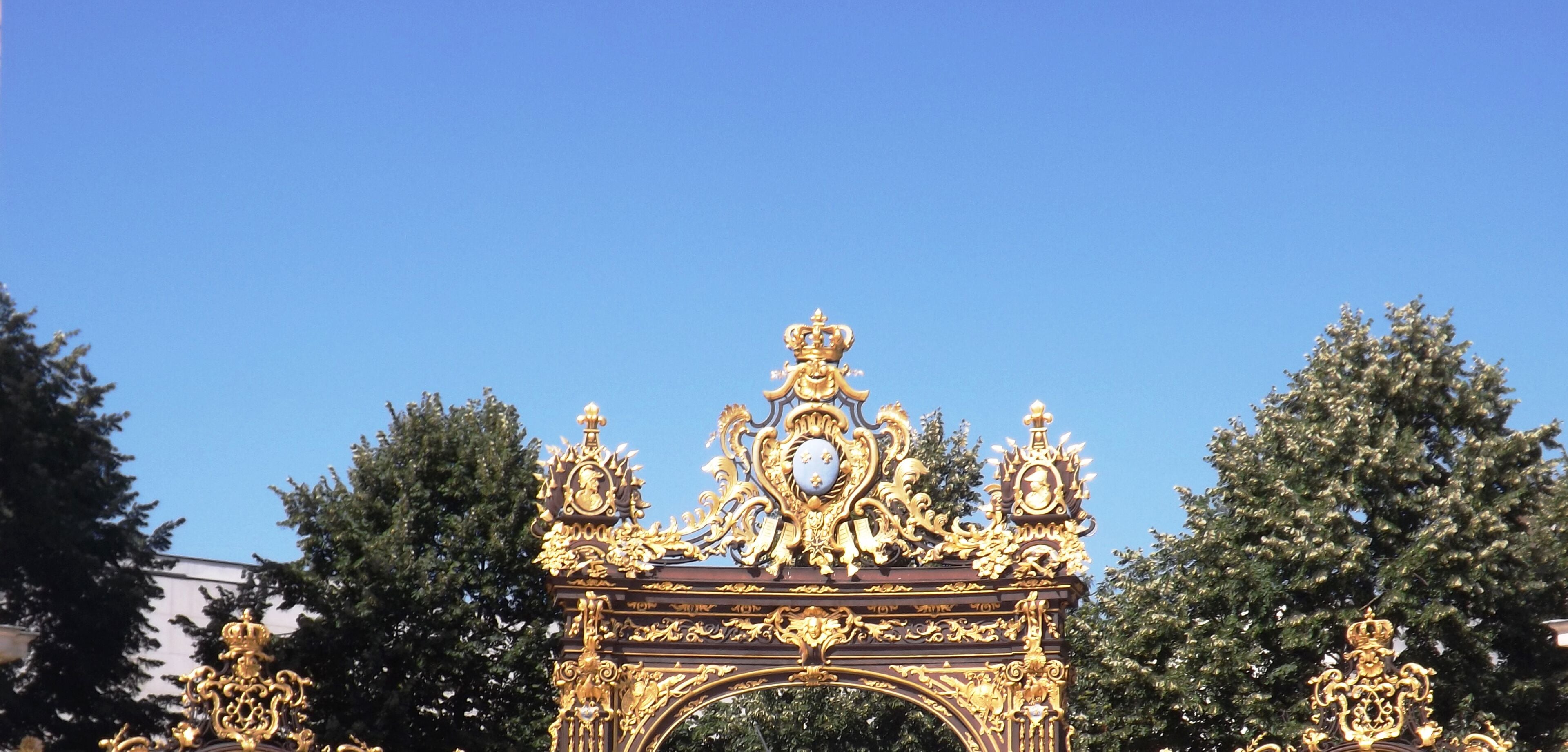 Una puerta dorada ornamentada con diseños intrincados y un reloj, rodeada de árboles.