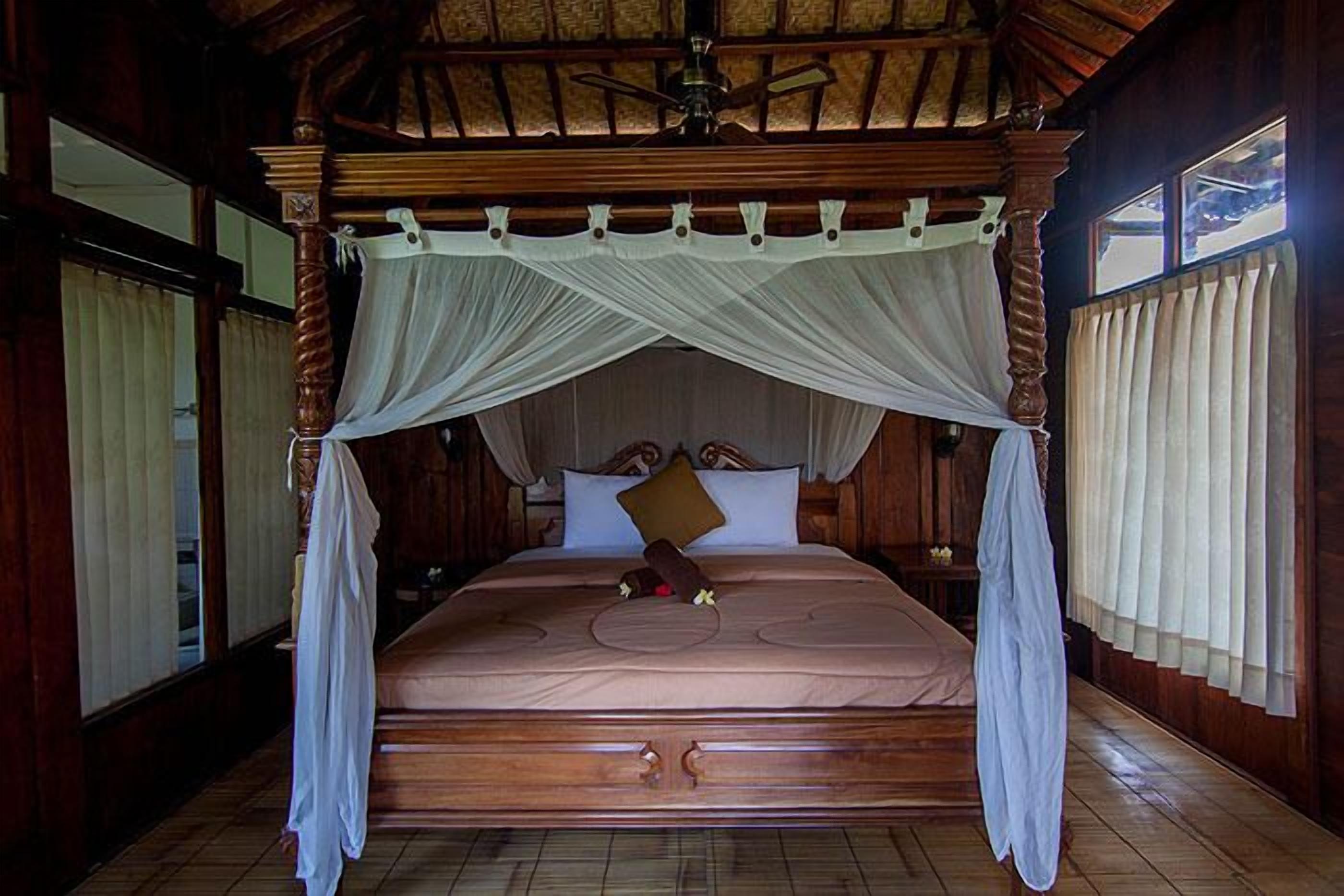 Una cama grande y ornamentada de madera con dosel, un ventilador de techo y cortinas en las ventanas.